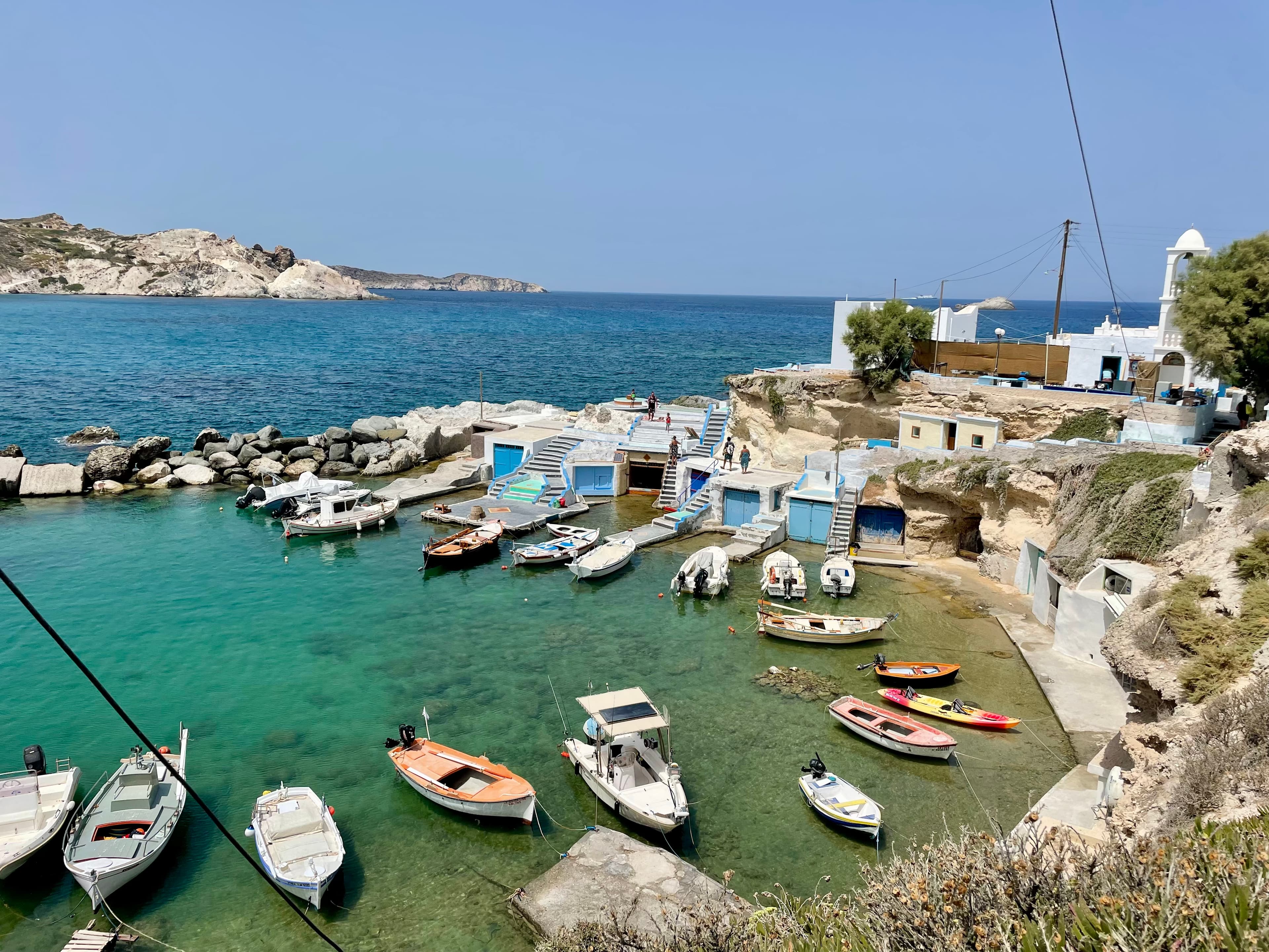 Small Milos fishing harbor — wooden boats moored in turquoise water beside painted syrmata boat houses, a whitewashed chapel on the headland