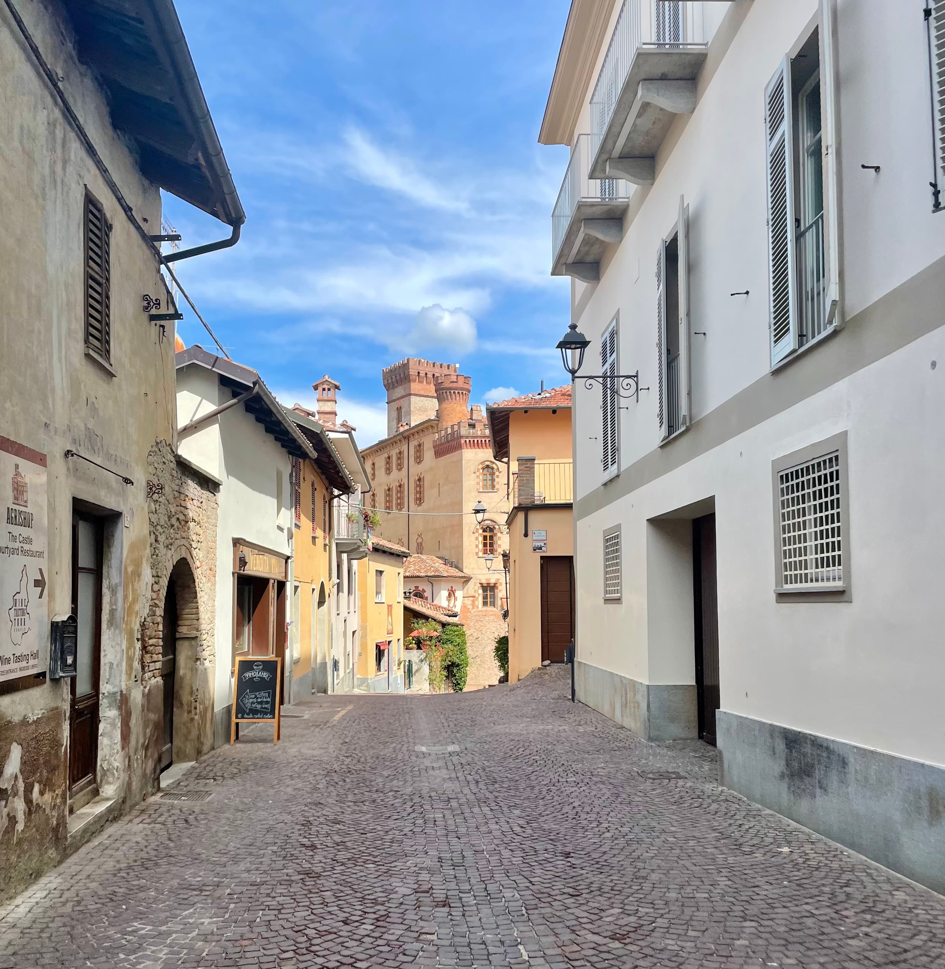 Cobbled Piedmont village street climbing toward a hilltop castle, shuttered stone houses lining either side
