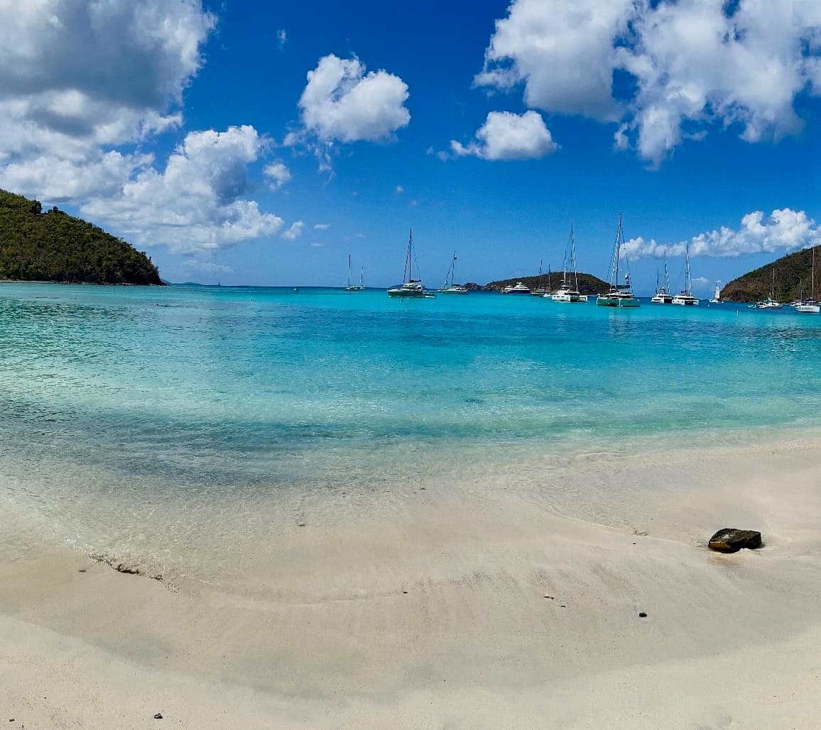 St. John North Shore beach — clear turquoise water, sailboats anchored in the bay, green headlands on either side, white sand in the foreground